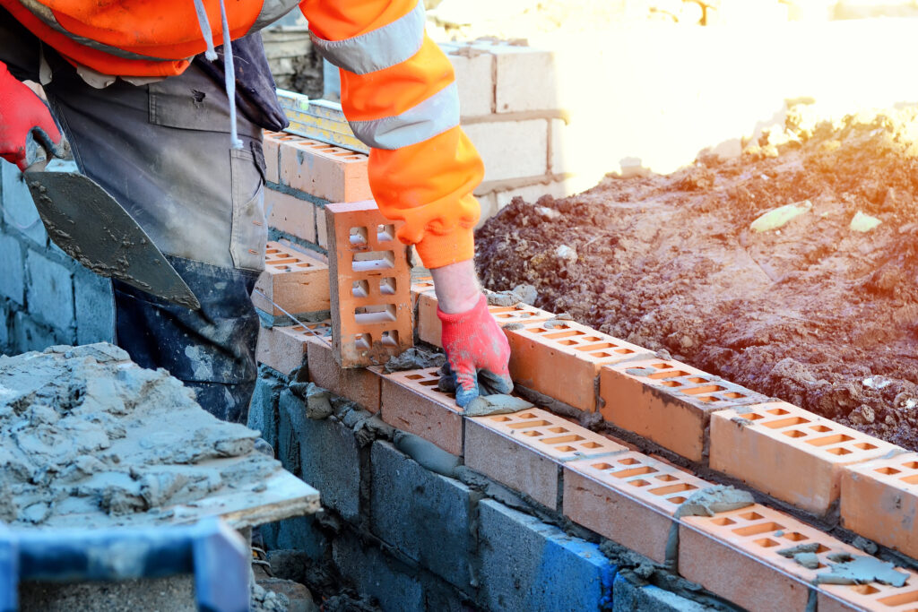 Industrial bricklayer laying bricks on cement mix on construction site. Fighting housing crisis by building more affordable houses concept